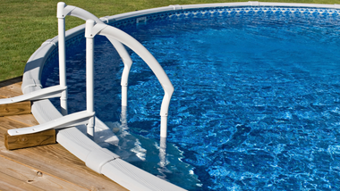 Above-ground swimming pool with white handrails leading into blue water, viewed from a wooden deck.