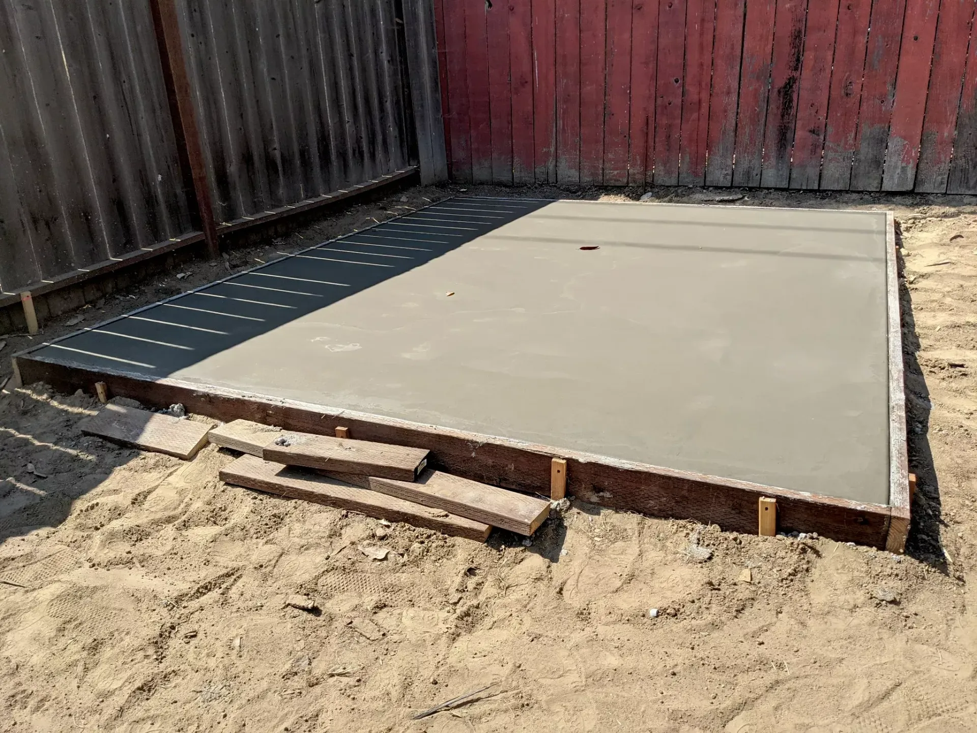 Freshly poured concrete slab in a backyard, surrounded by wooden framing and a red fence.