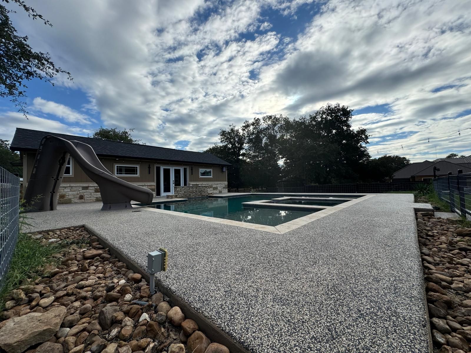 Pool with slide, connected walkway, beige house, and cloudy sky. Pebbles in foreground.