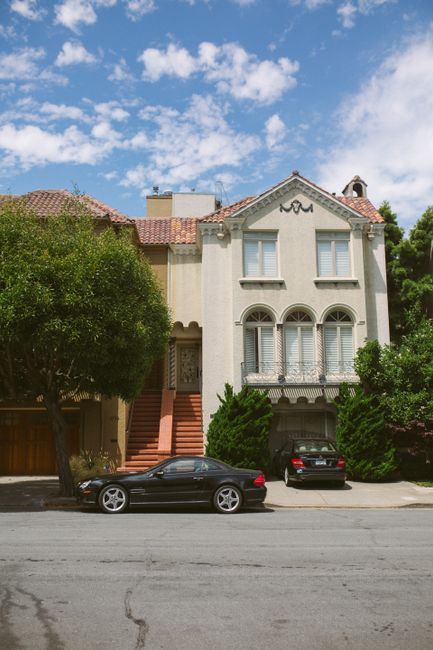 Street view of a cream-colored townhouse with parked cars and trees under a partly cloudy sky