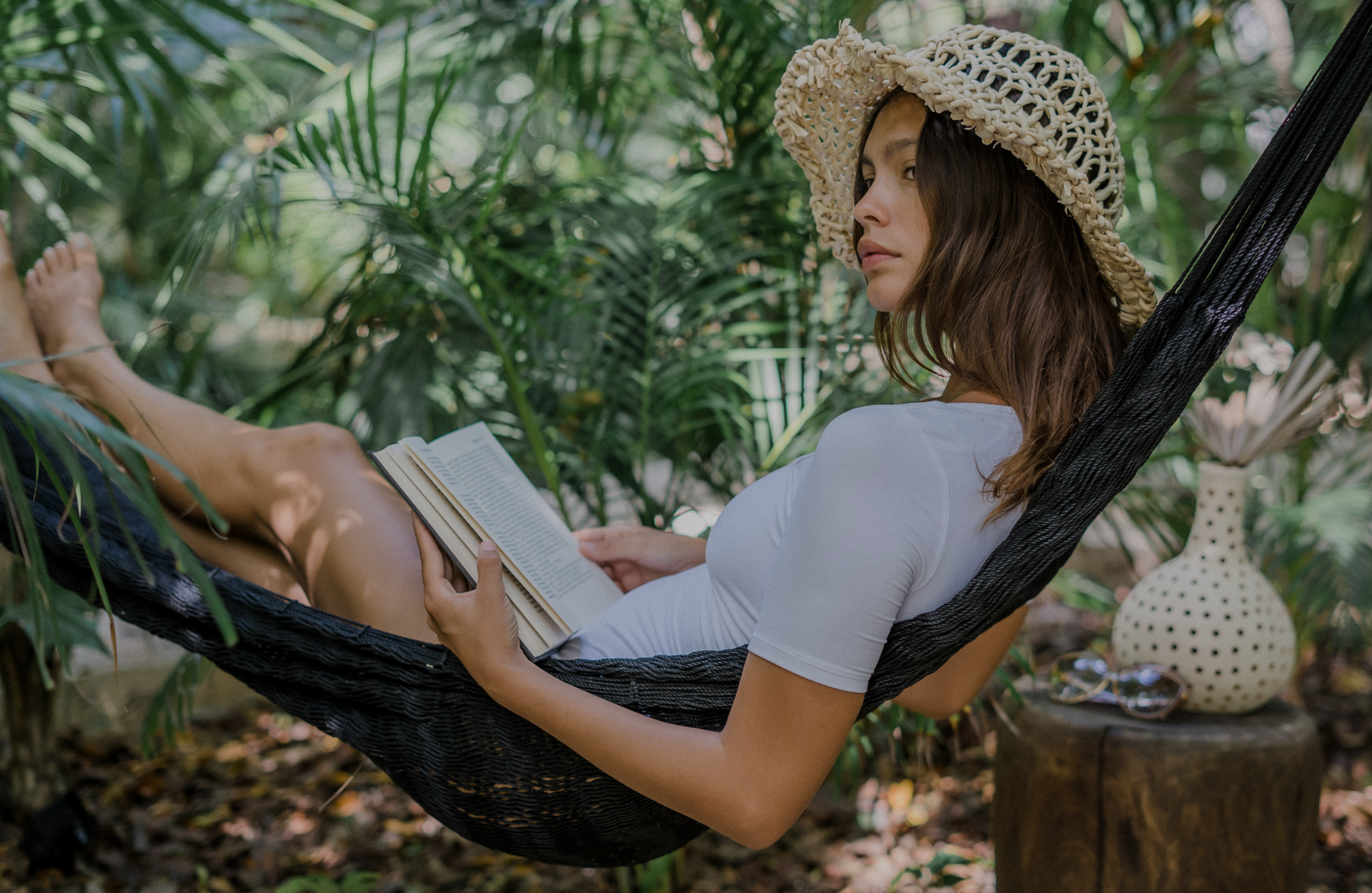 Woman wearing a sun hat and white dress reads a book while relaxing in a hammock outdoors.