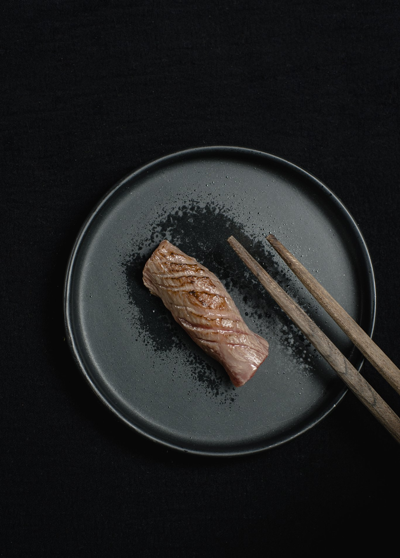 A piece of sushi on a black plate with chopsticks.