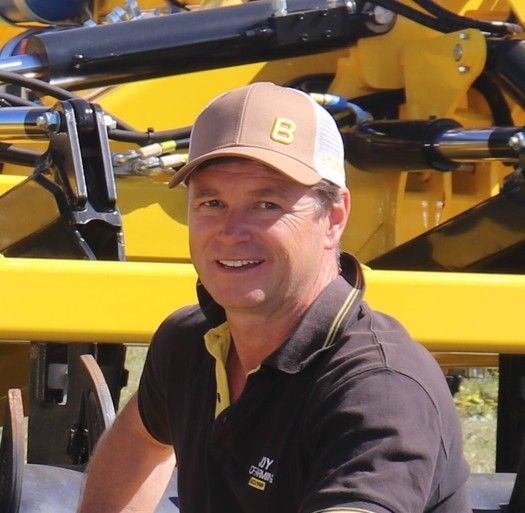 Man wearing a brown cap and polo shirt smiles, posing in front of yellow farm equipment.