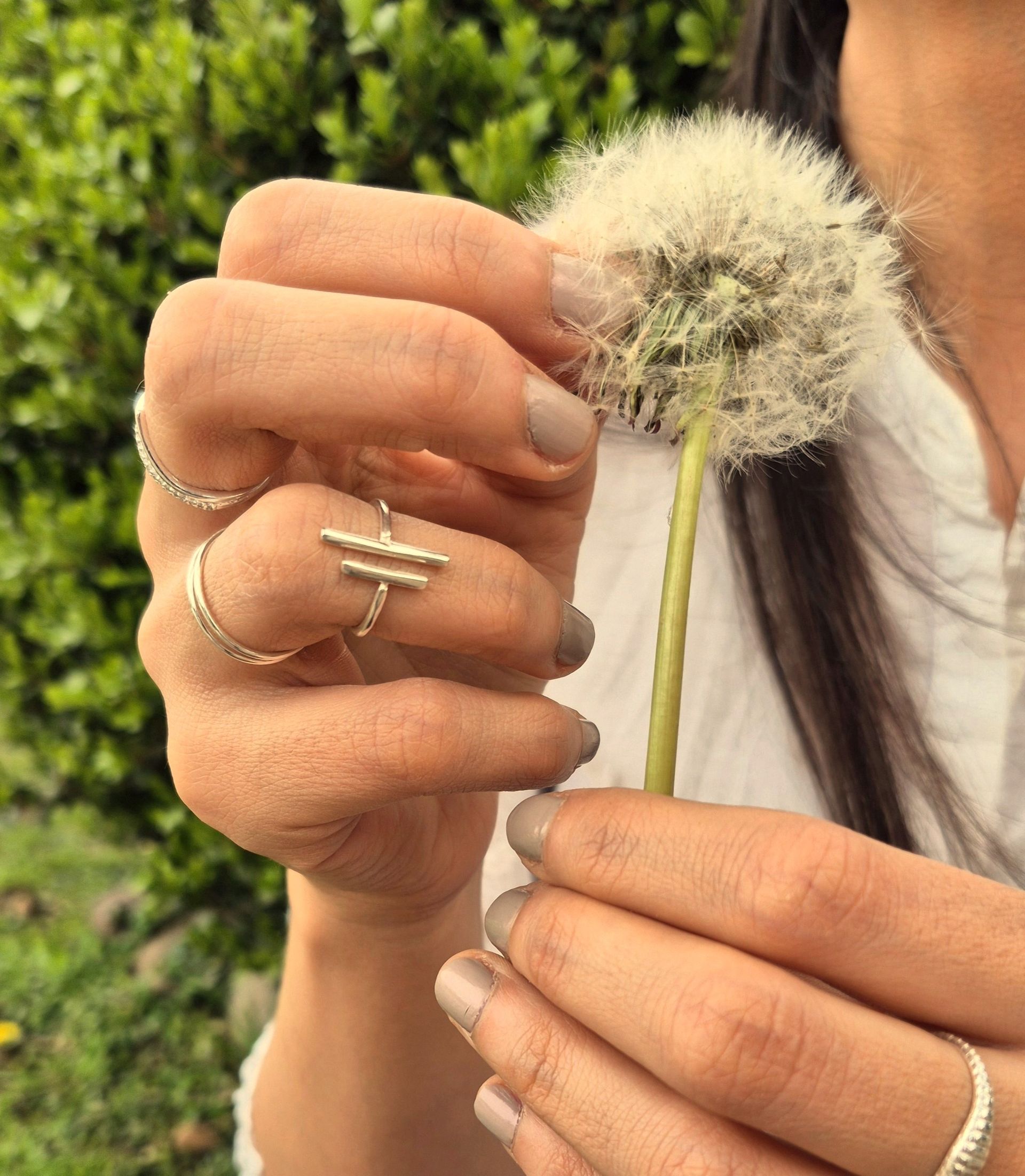 Persona sosteniendo una cabeza de semilla de diente de león con los dedos adornados con anillos, al aire libre.