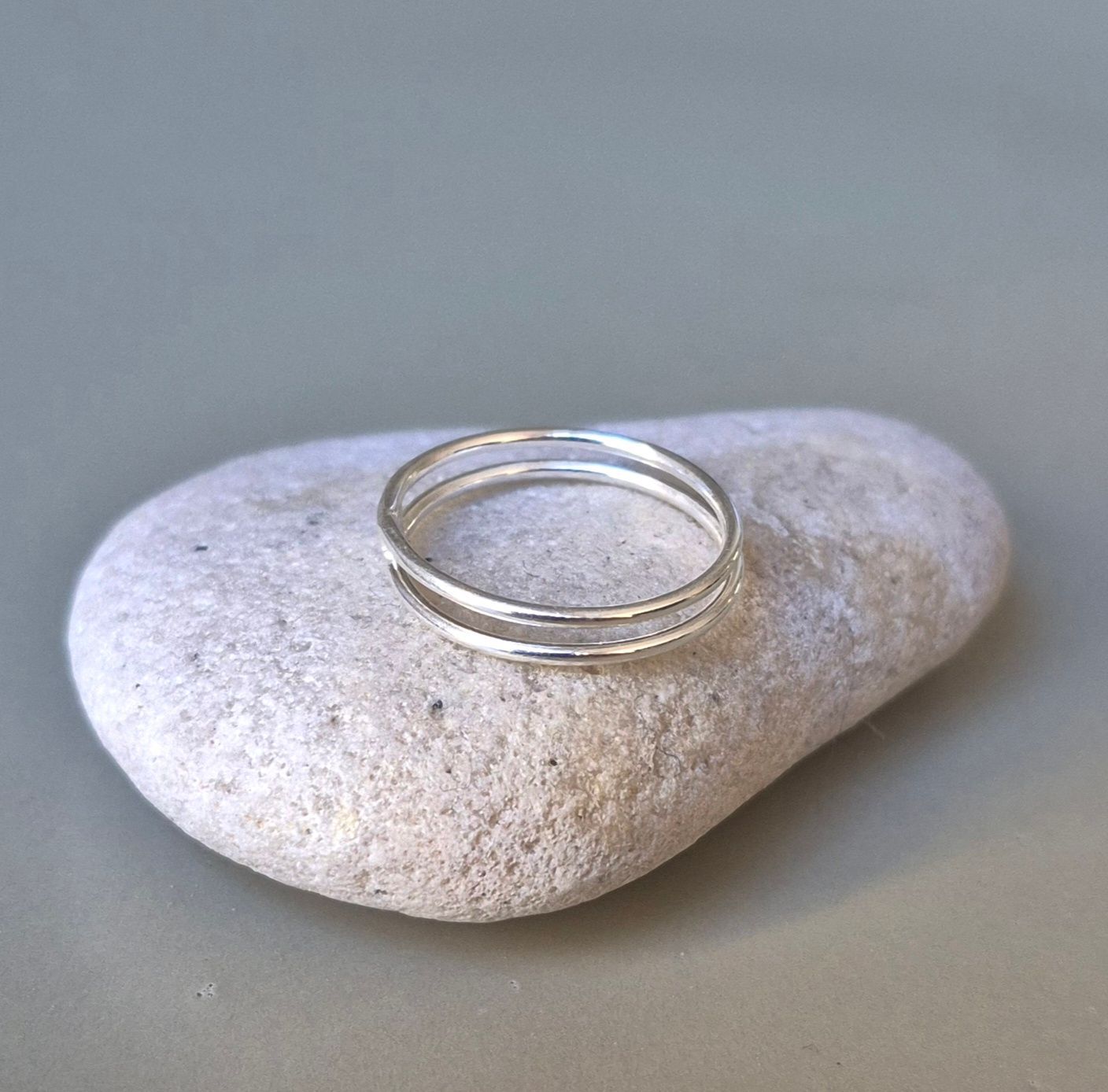 Anillo de plata de doble banda con una piedra texturizada de color gris claro sobre un fondo neutro.