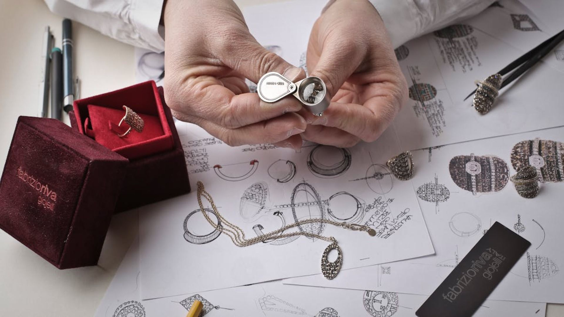 Manos que sostienen un pequeño anillo medallón de plata, rodeadas de bocetos de joyas y una caja para anillos.