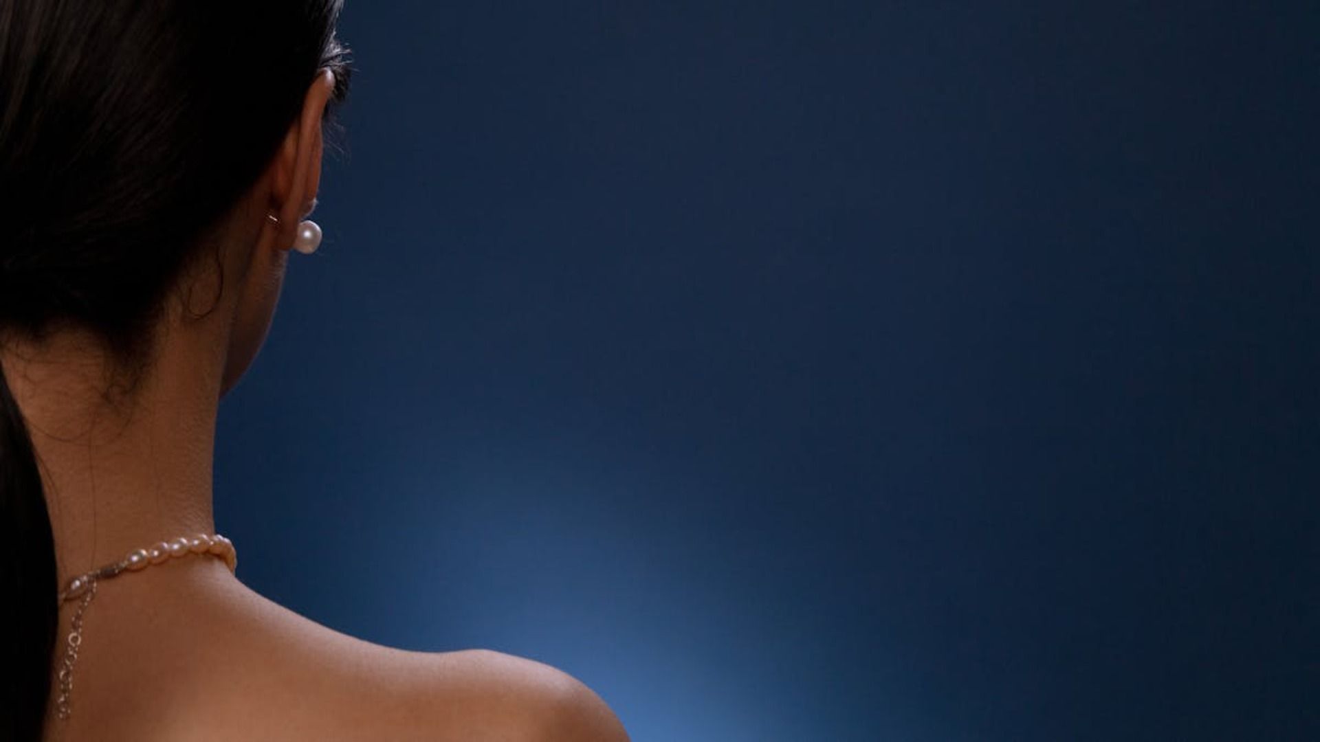 Espalda y hombro de una mujer, con pendientes y collar de perlas, sobre un fondo azul oscuro.