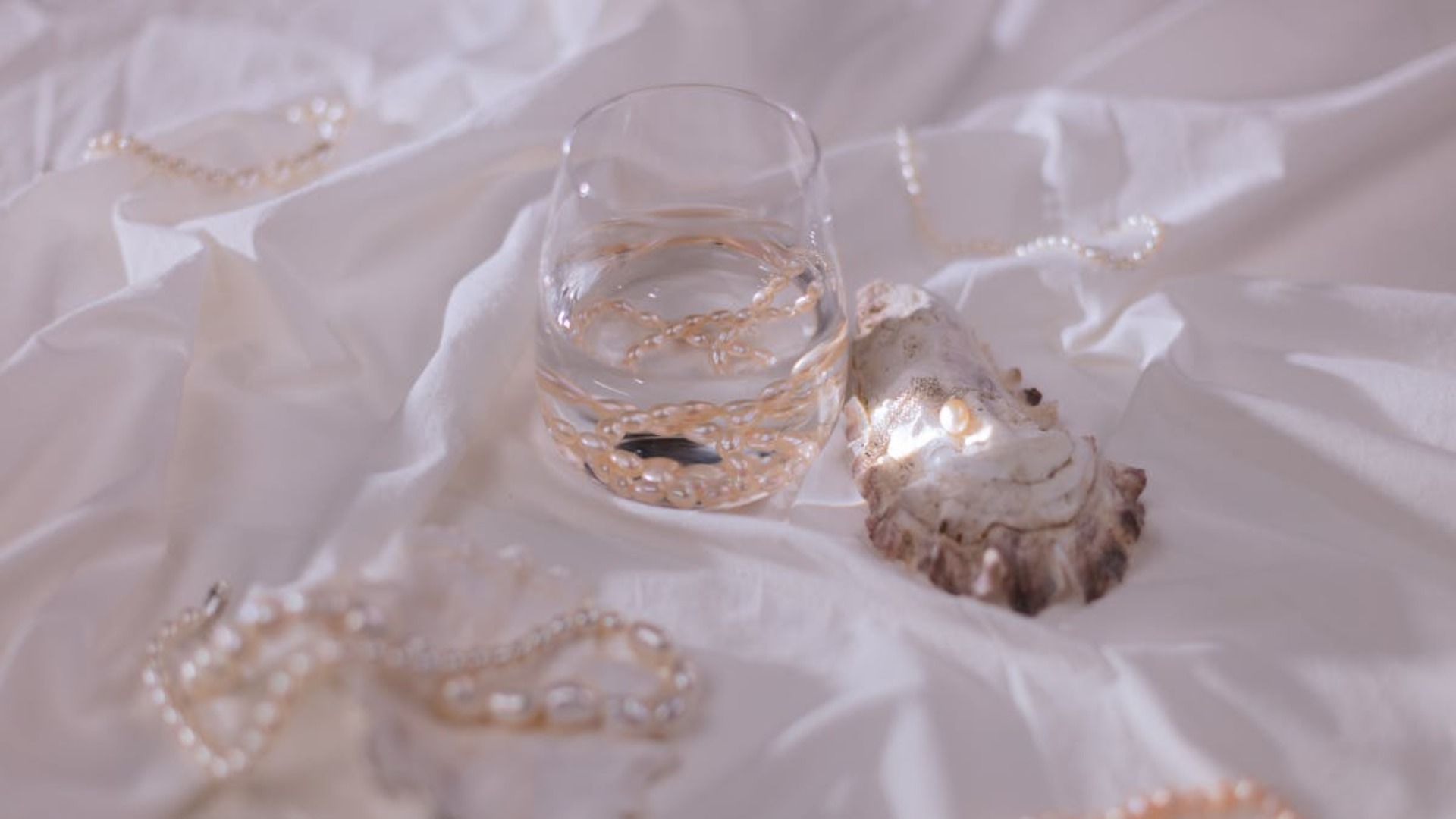 Vaso de agua con joyas doradas, concha de ostra y tela blanca drapeada.