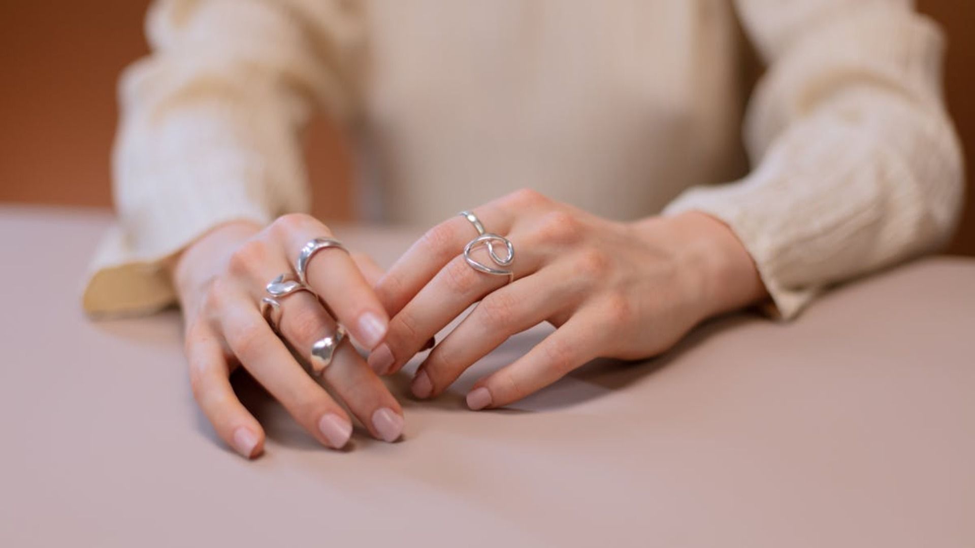 Manos con anillos descansando sobre una superficie de color claro, vestidas con una blusa color crema.