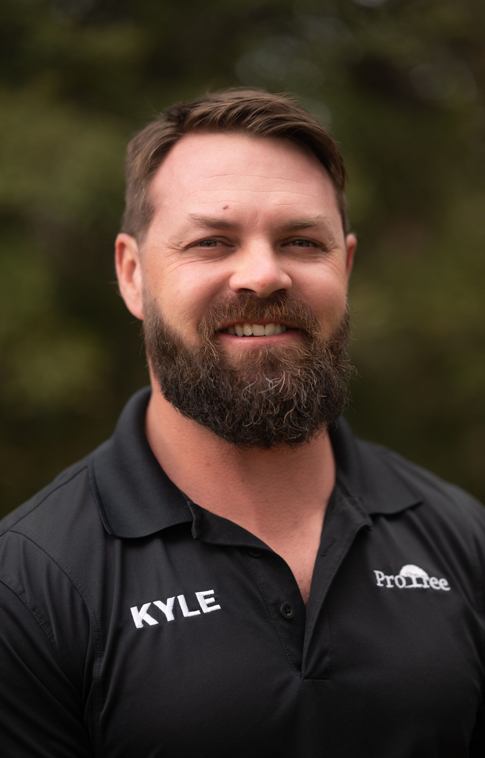 Smiling man in a black polo shirt with name “Kyle” on it, standing outdoors.