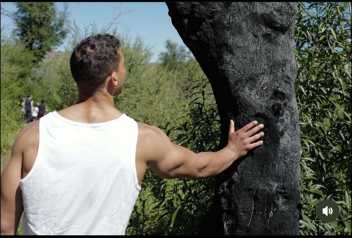 A man in a white tank top is standing next to a tree