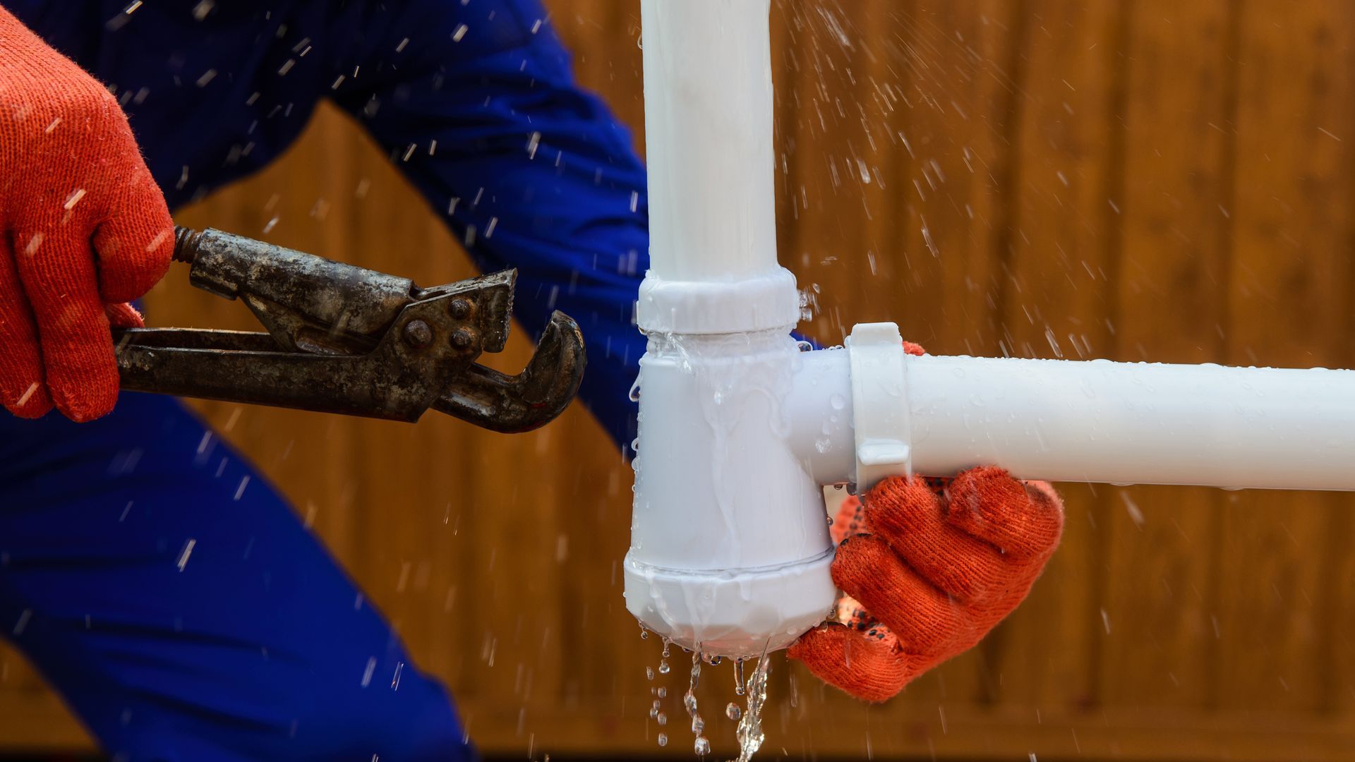 Plumber in blue coveralls and red gloves fixing a leaking white pipe with water spraying.