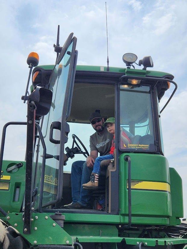 Two people inside a green tractor cab, one standing on the steps against a cloudy sky.