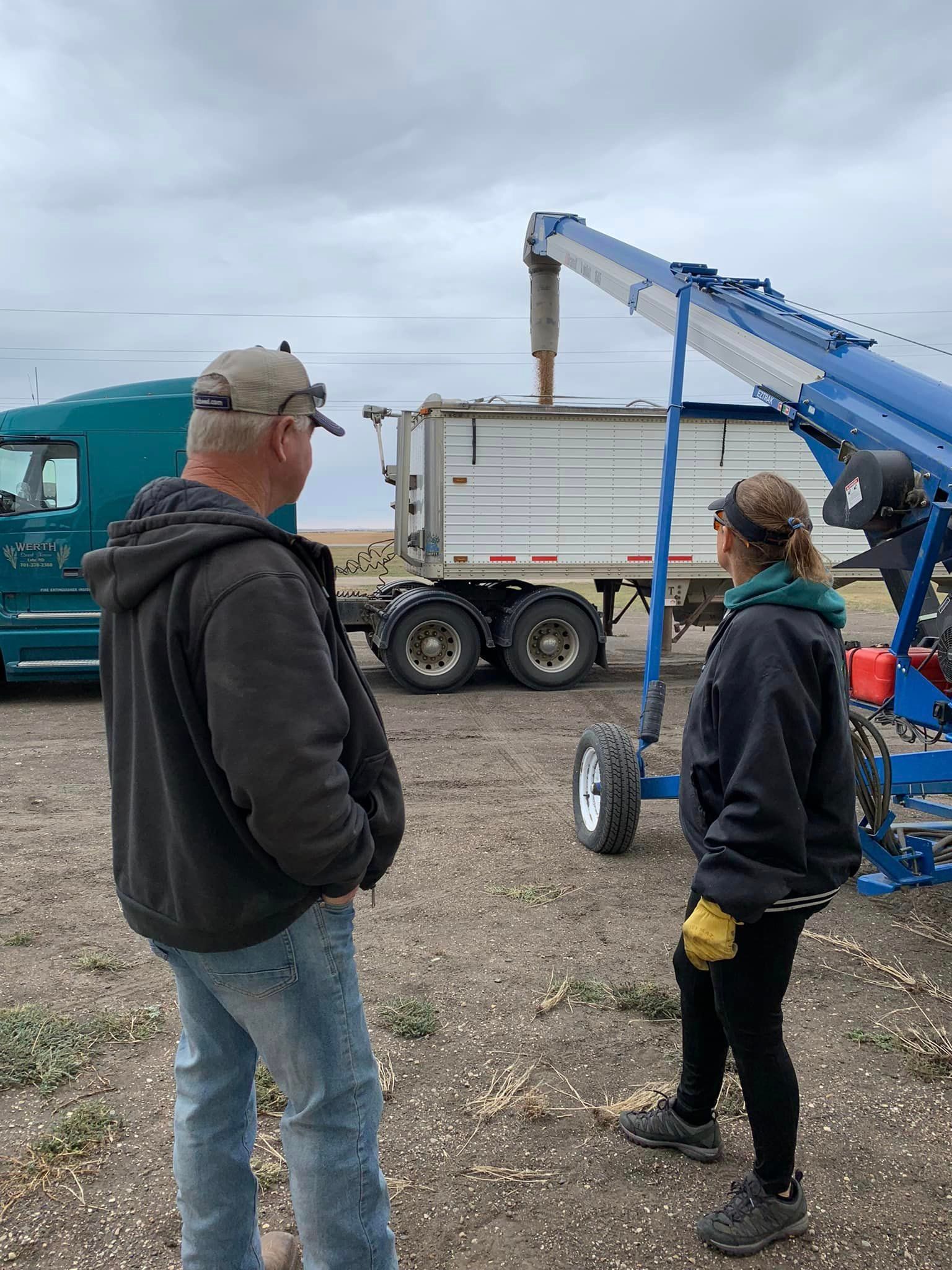 Two people standing beside a white dump truck and blue boom lift in a gravel lot