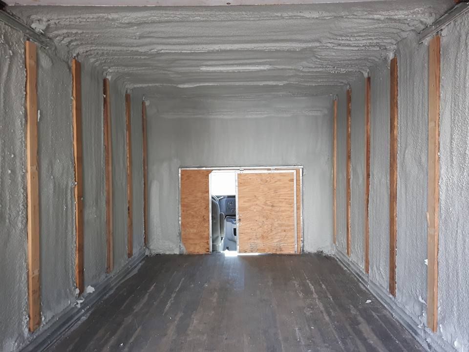 Intérieur d'un conteneur isolé avec de la mousse grise. Des poutres en bois bordent les murs. Une porte se trouve au fond.