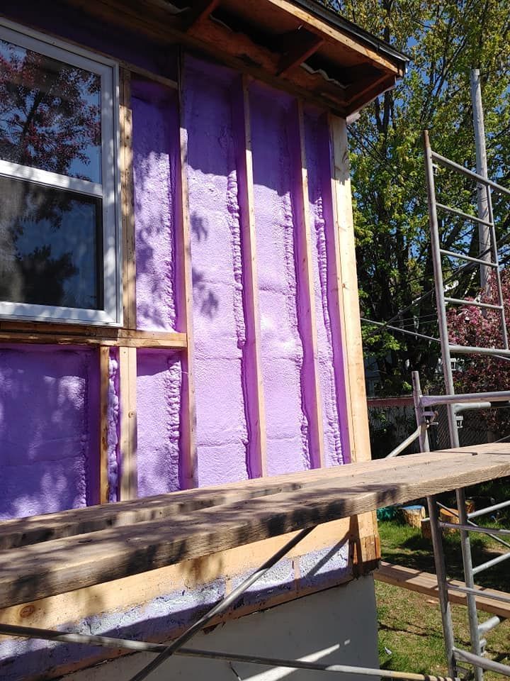 Extérieur de la maison en construction, montrant une isolation en mousse projetée rose entre les montants en bois. Un échafaudage est présent.