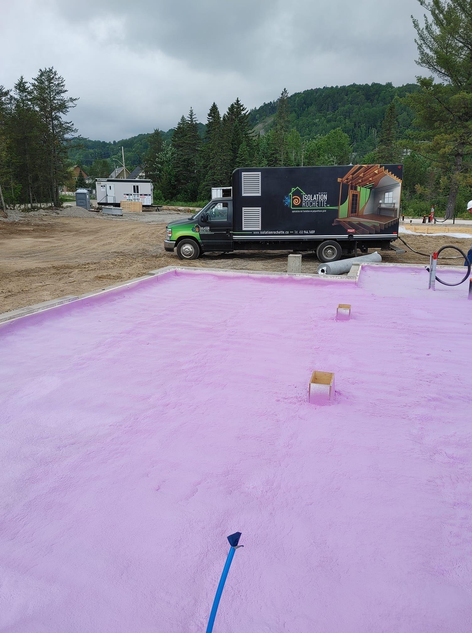 Camion vert avec logo garé près d'un chantier de construction avec une fondation en mousse rose par une journée nuageuse.