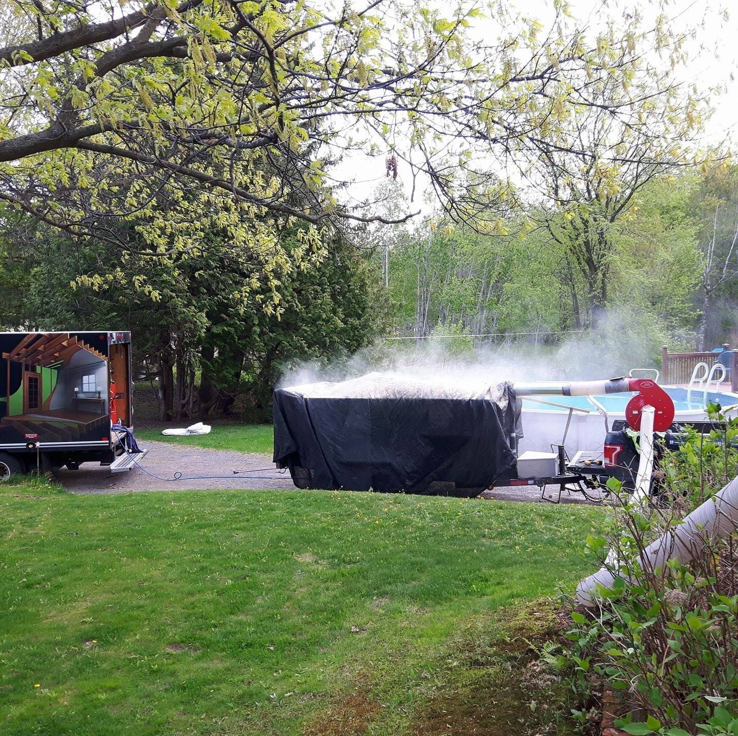 Bain à remous fumant en extérieur. Grand réservoir recouvert d'une bâche noire. Un camion et des arbres verts à proximité.