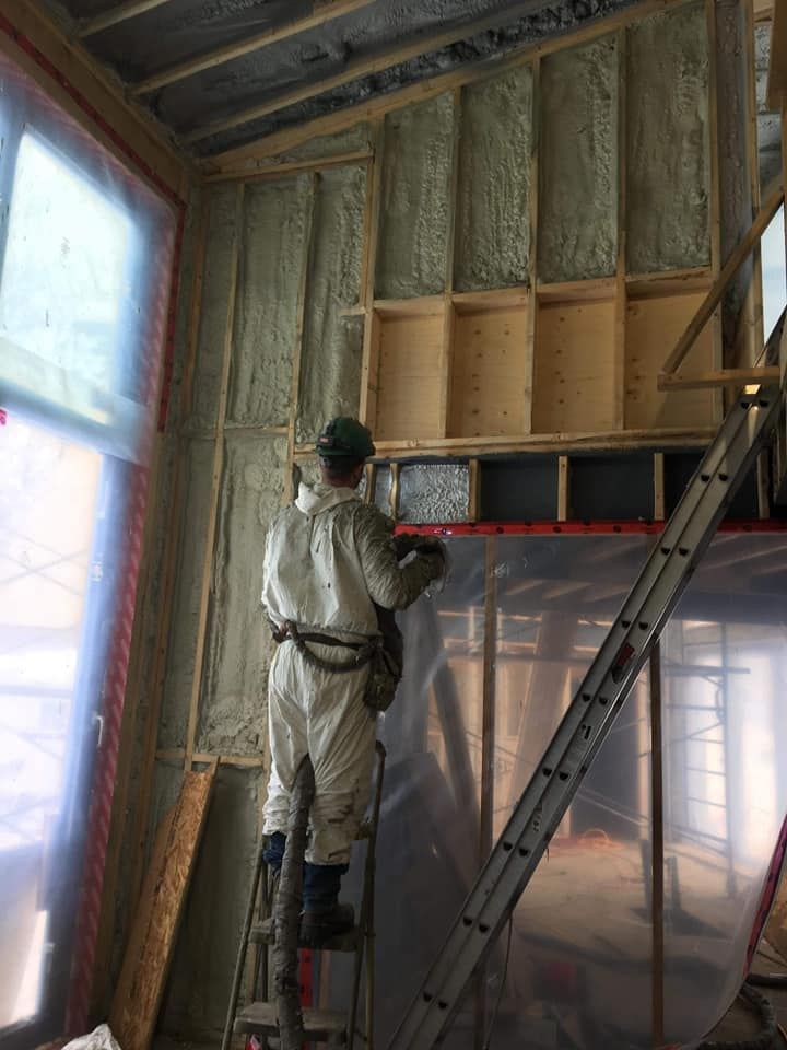Une personne en combinaison de protection pulvérise de l'isolant sur un mur intérieur à l'aide d'une échelle.