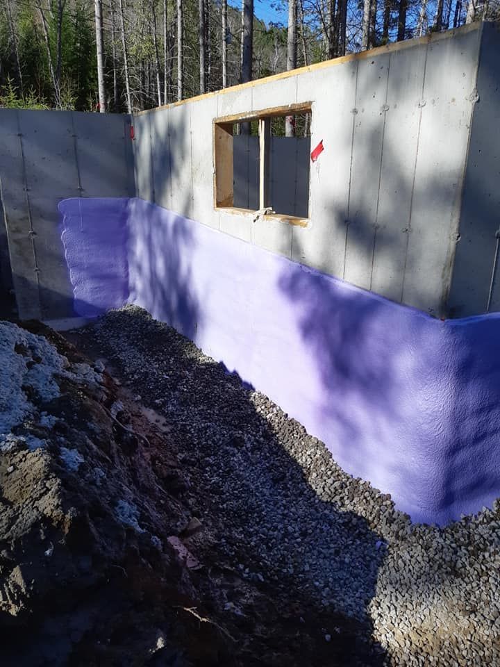 Fondation en béton avec isolation violette, gravier et ouverture de fenêtre dans une zone boisée.