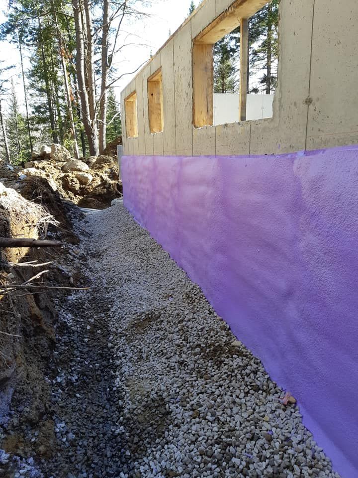 Un mur de fondation avec une imperméabilisation violette, du gravier et une tranchée ; à l'extérieur, des arbres.