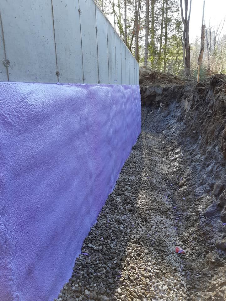 Isolation en mousse violette sur un mur de fondation en béton, une tranchée de drainage en gravier, un talus de terre et un arrière-plan forestier.