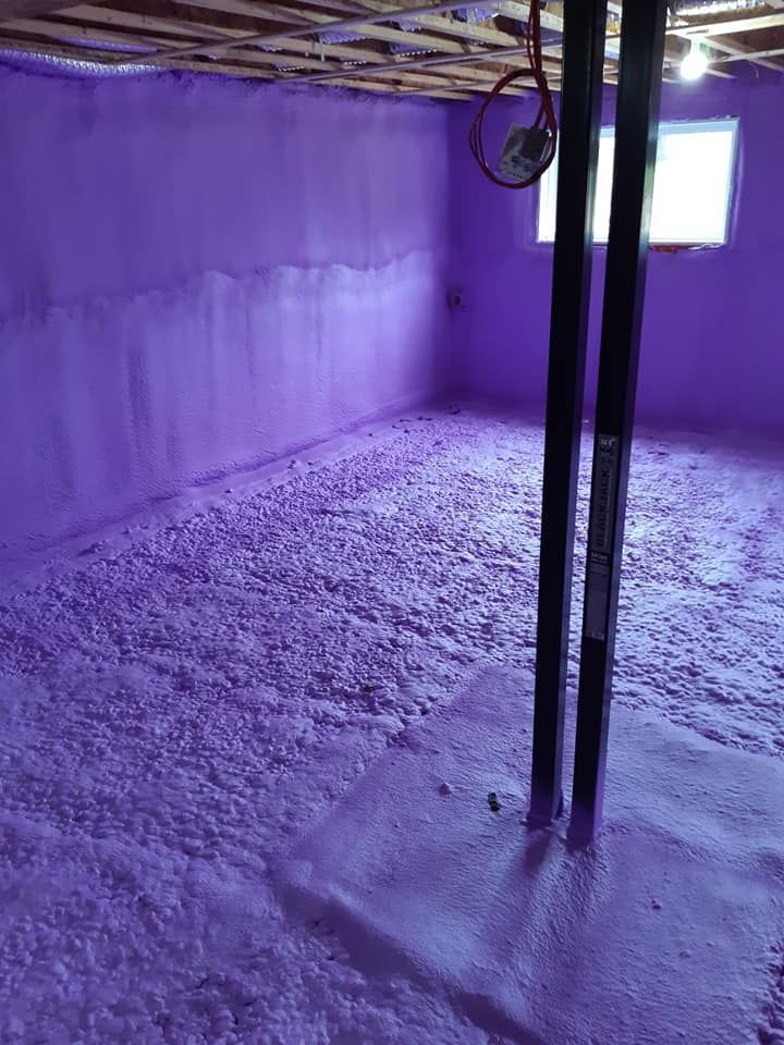 Une mousse isolante violette pulvérisée recouvre les murs et le sol d'un sous-sol. Une poutre de soutien est visible.