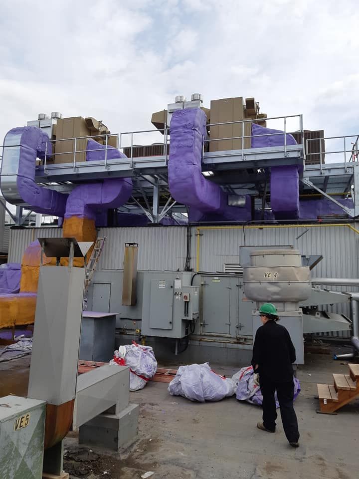 Une installation industrielle avec des tuyaux isolés violets, un ouvrier et des sacs ; des machines grises et une structure métallique ; un ciel couvert.