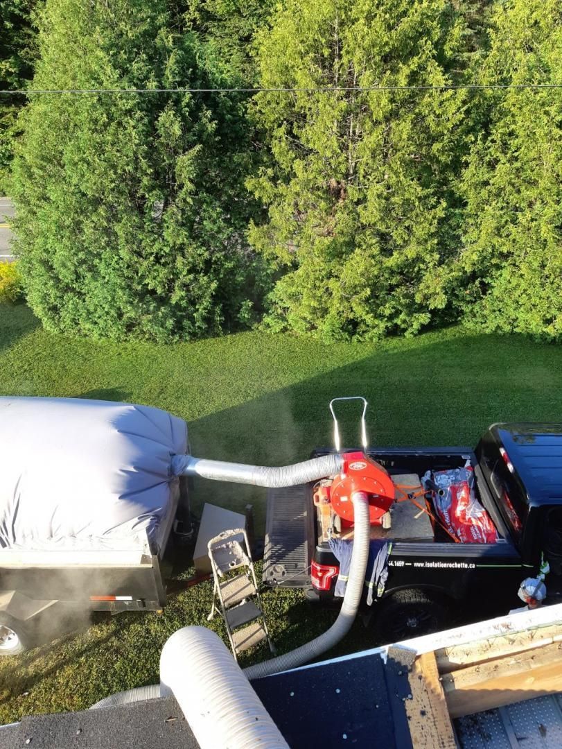 Camion avec équipement, tuyau raccordé au toit, probablement pour la pose d'isolant. Herbe verte et arbres en arrière-plan.
