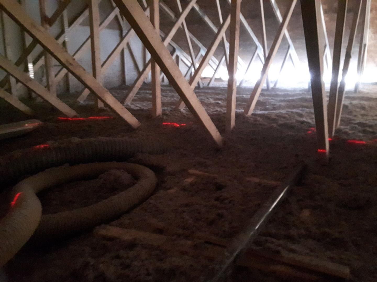 Vue en contre-plongée d'un grenier avec des fermes en bois recouvrant un isolant en vrac, montrant de faibles lignes rouges de nivellement laser.