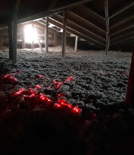 Vue d'un grenier sombre, dont le sol est recouvert d'une épaisse isolation grise et où brillent des lumières décoratives rouges au premier plan.