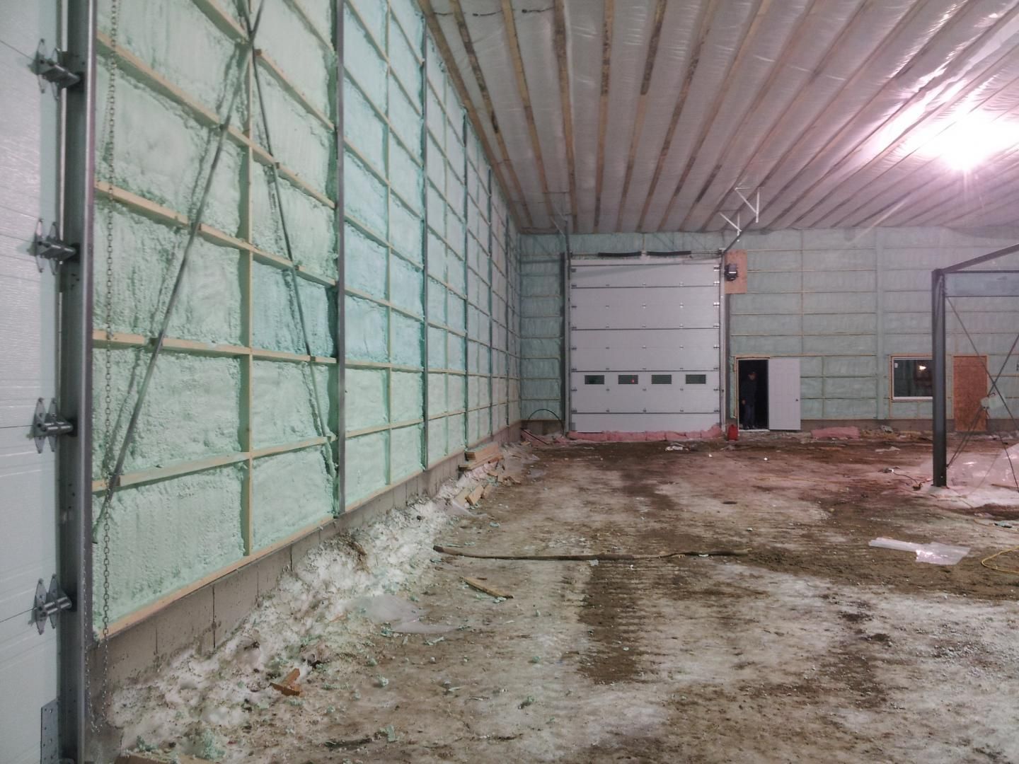Intérieur d'un bâtiment avec isolation en mousse sur les murs, porte de garage fermée et sol en béton sale.