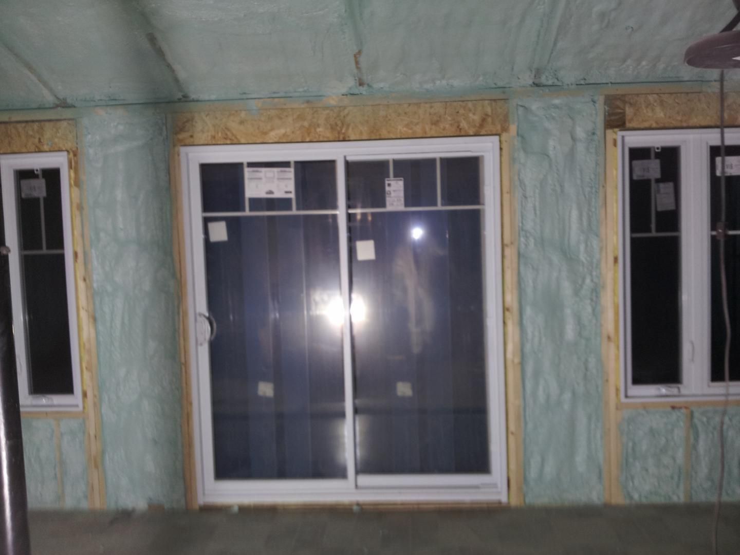 Vue intérieure d'une pièce en construction, montrant une porte coulissante en verre et des fenêtres encadrées de bois, isolées avec de la mousse bleue.