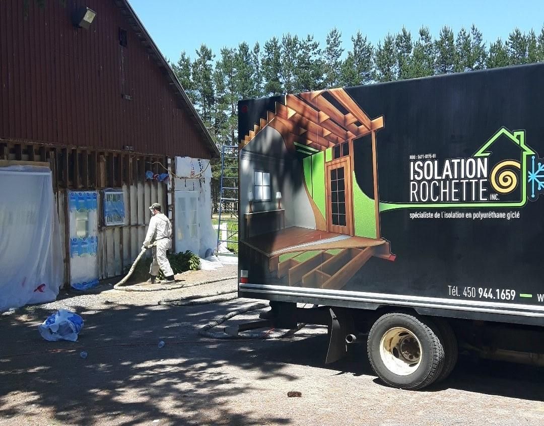 Un ouvrier portant un équipement de protection applique de l'isolant sur le mur d'un bâtiment à côté d'un camion-citerne de marque Isolation Rochette.