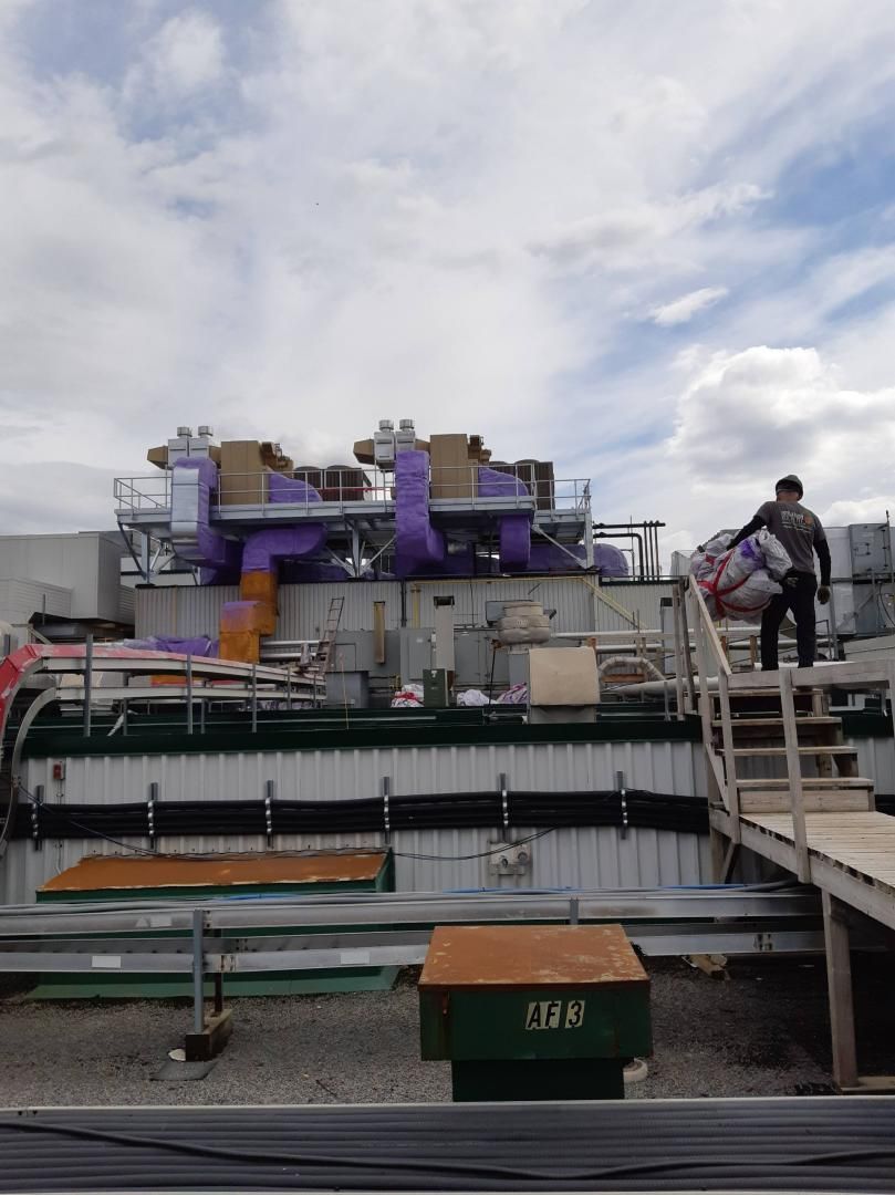 Une personne monte un escalier en bois vers un grand équipement industriel violet et gris, à l'extérieur sous un ciel nuageux.