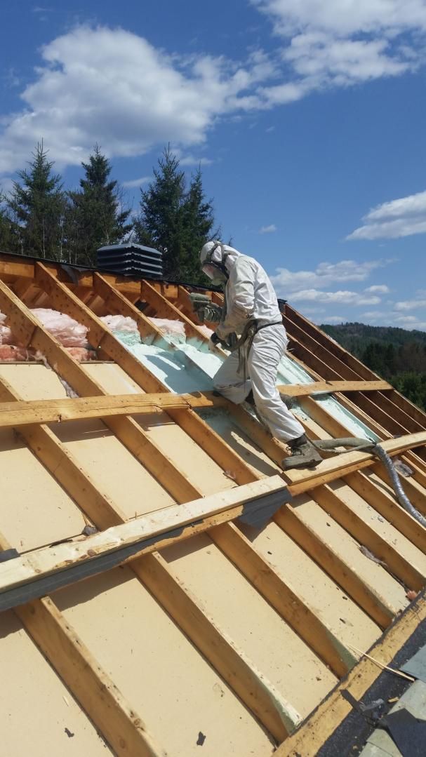 Un ouvrier en combinaison de protection pulvérise de l'isolant sur un toit sous un ciel bleu.