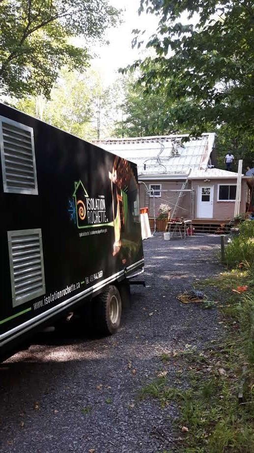Camion noir stationné près d'une maison avec des ouvriers sur le toit ; arbres en arrière-plan.