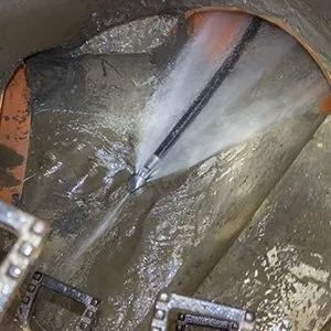 A high-pressure jetting nozzle sprays water to clean a dark, concrete underground pipe or sewer maintenance hole.