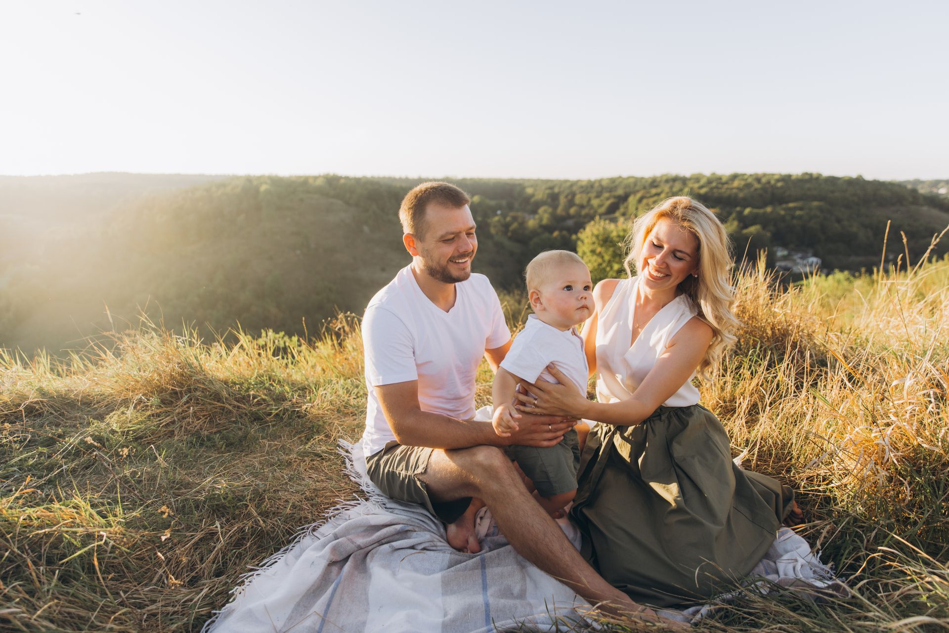 A family is sitting on a blanket in a field holding a baby.