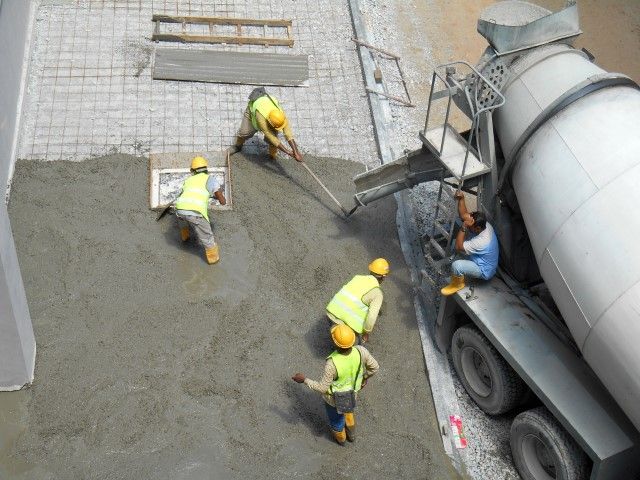 Construction workers pouring concrete from a truck, spreading it on a surface with rebar, wearing safety vests and hard hats.