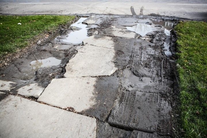 Damaged sidewalk with broken concrete and puddles next to a grassy area and road.
