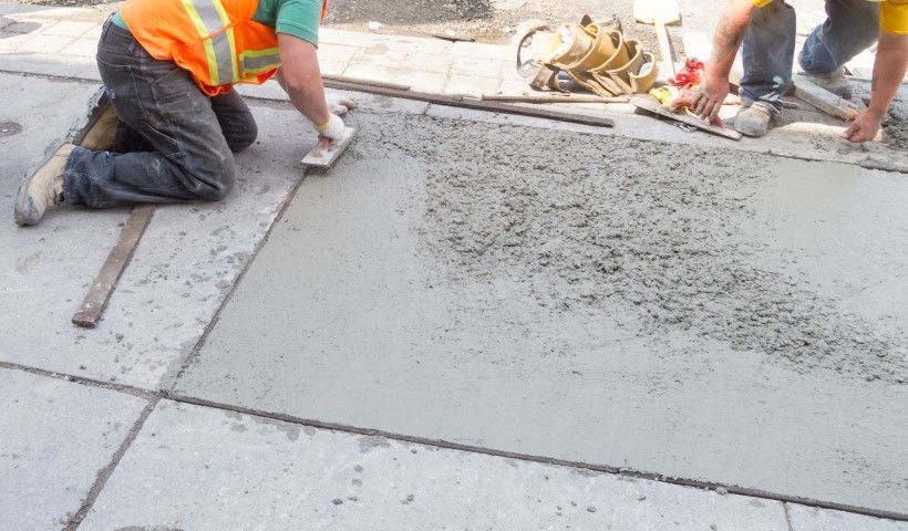 Workers smoothing wet concrete sidewalk with tools, wearing safety vests.