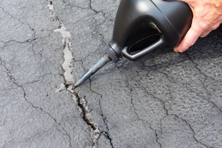 Person pouring sealant from a black jug into a crack in asphalt pavement.