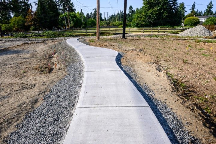 Concrete pathway winding through a grassy area, gravel on the edges, trees in the background.
