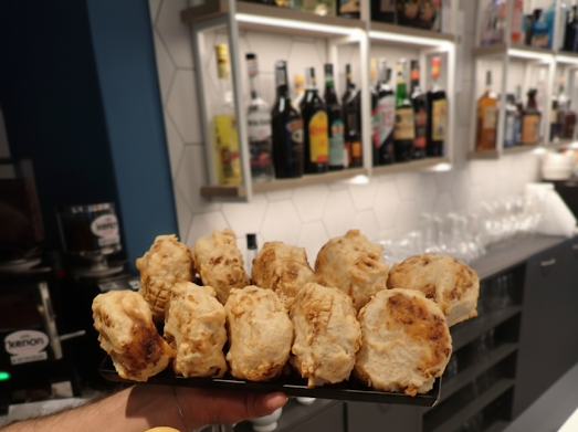 Persona che regge un vassoio di cibo fritto dorato in un bar, con bottiglie di liquore sugli scaffali sullo sfondo.
