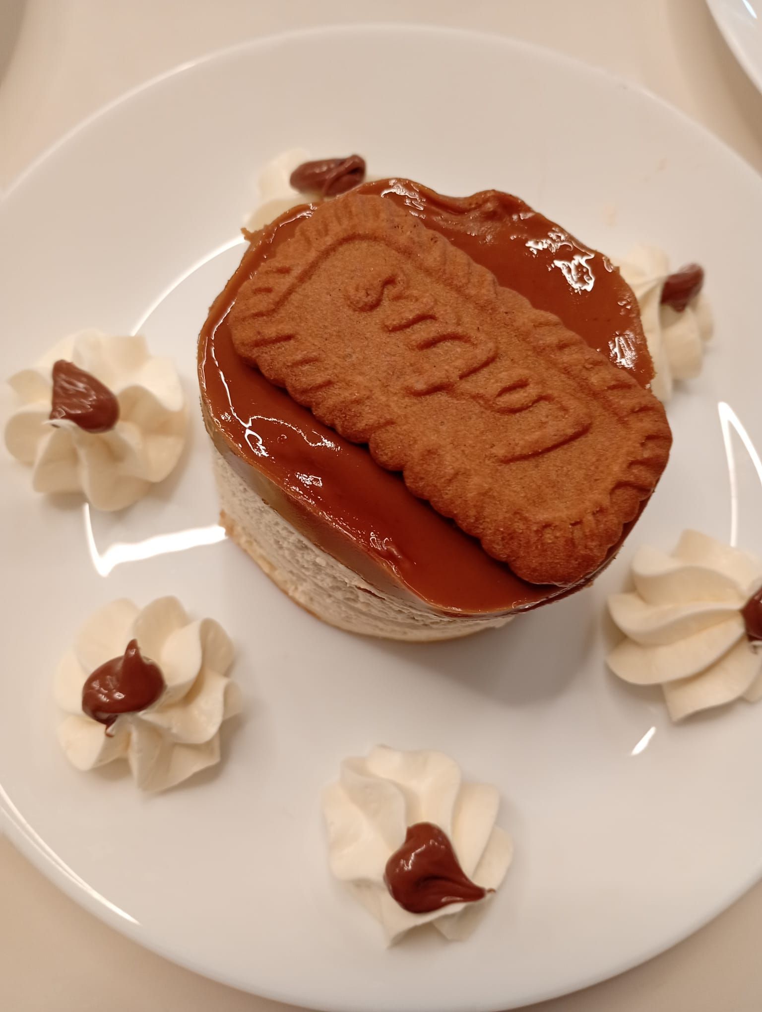 Dessert su un piatto bianco: una torta guarnita con caramello e un biscotto Lotus, circondata da rosette di panna montata.