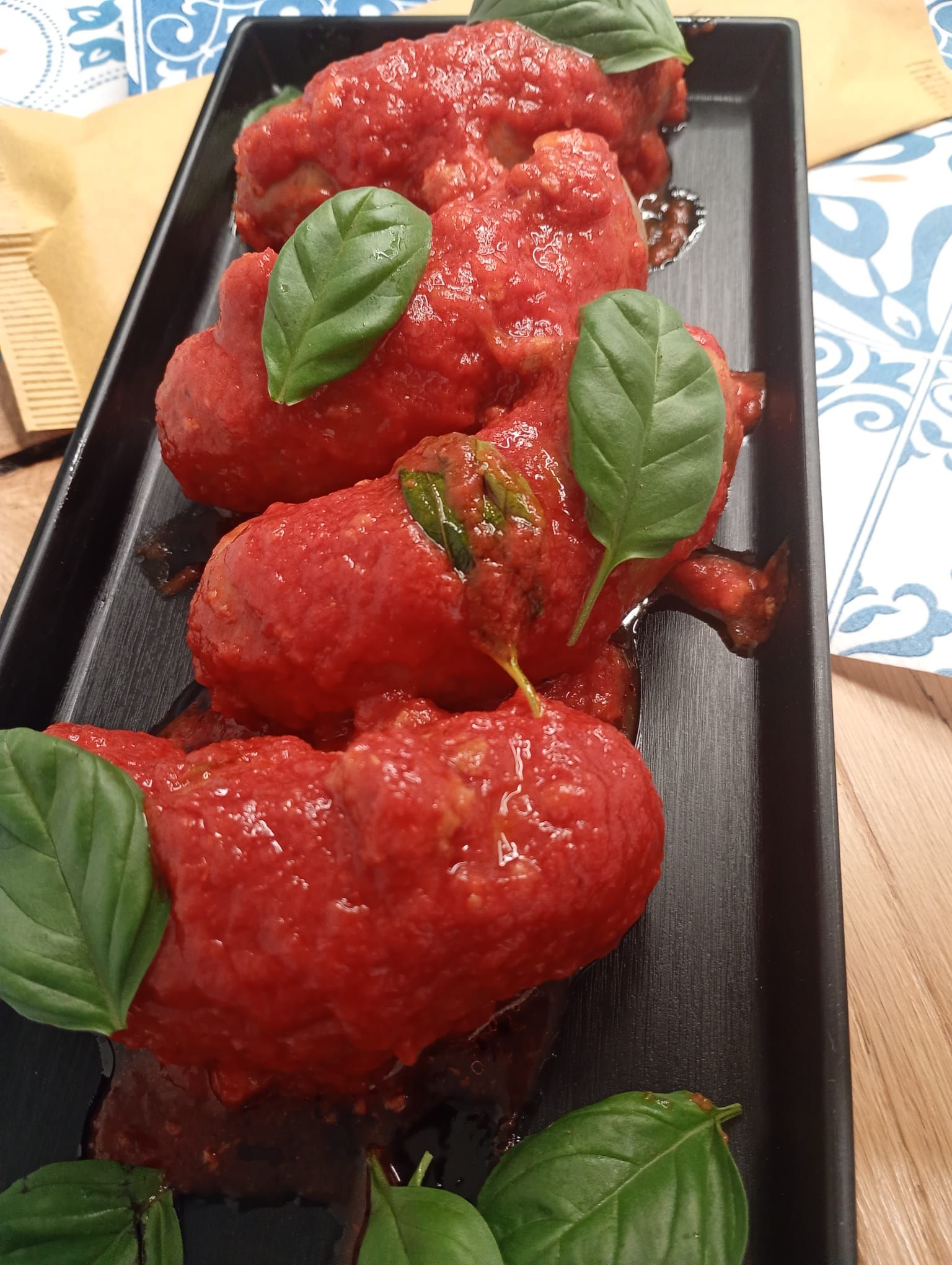 Polpette in salsa rossa, guarnite con foglie di basilico, su un piatto rettangolare nero.