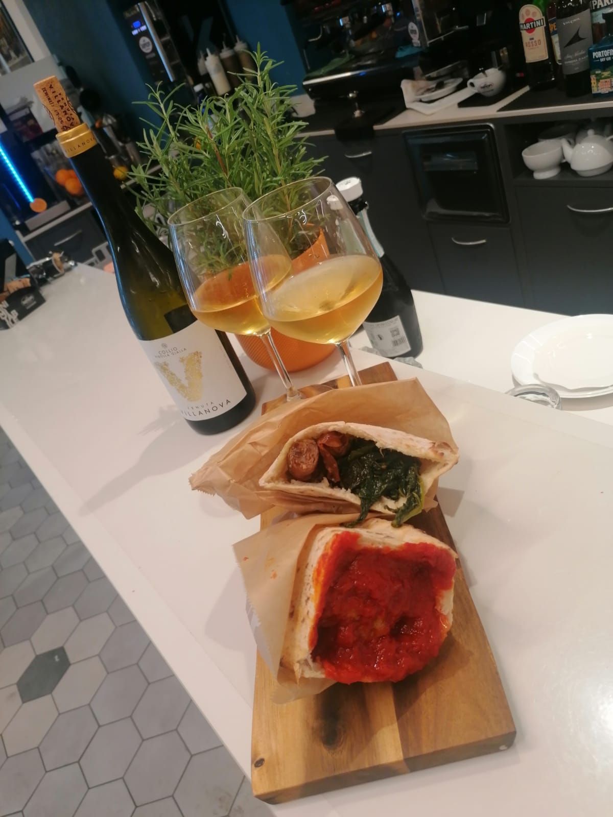 Un tagliere di legno con pizza e panini, accanto a bicchieri di vino e bottiglie su un bancone bianco.