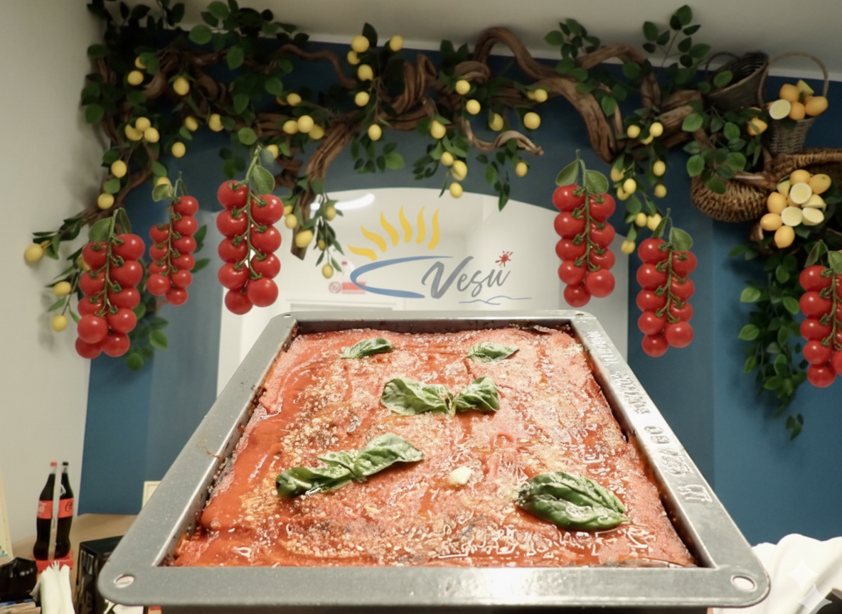 Grande teglia rettangolare di pasta al forno con salsa di pomodoro, guarnita con basilico. Esposta su un bancone decorato con pomodori e limoni.