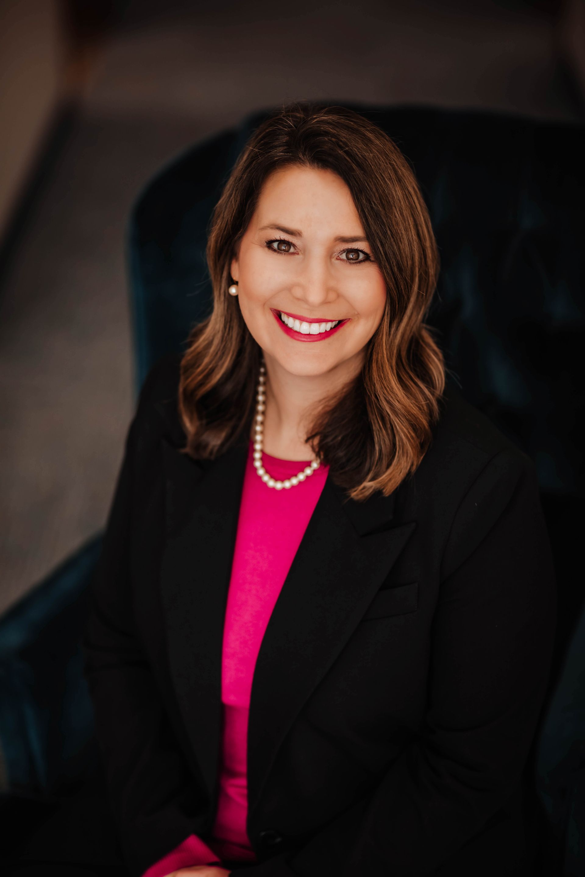 A woman is sitting in a chair wearing a black jacket and a pink shirt.